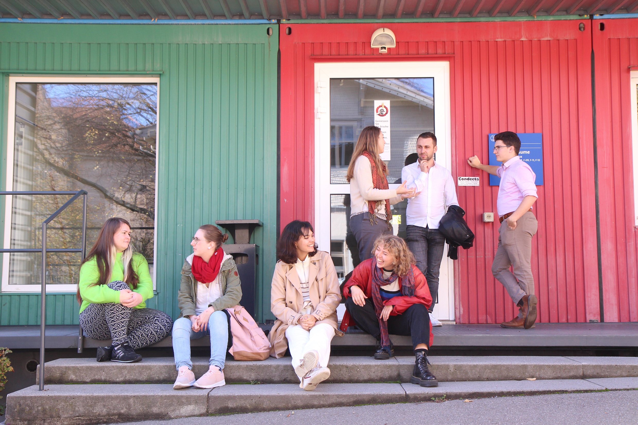 Das Bild zeigt eine Gruppe von Studierenden, die vor einem grün-roten Gebäude der Universität lebhaft diskutieren.
