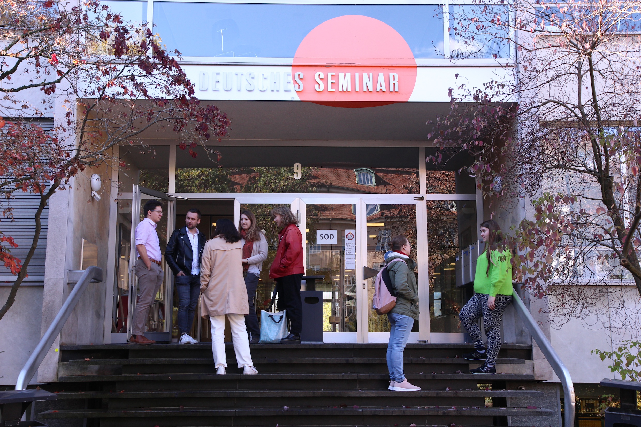 Das Bild zeigt eine Gruppe von Studierenden auf der Treppe vor dem Deutschen Seminar, sie sind ins Gespräch vertieft.